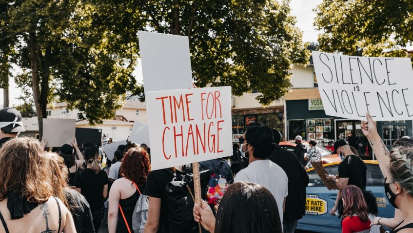 Diverse crowd protesting on the street. Protest signs say, "time for change" and "silence is violence."