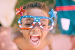 Child with mouth wide open and eyes closed wearing US flag-themed glasses with one big red star on one side, and one big blue star on the other side.