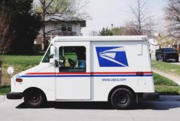 USPS mail truck parked on the street, with a house in the background. The mail carrier is seated inside the vehicle.