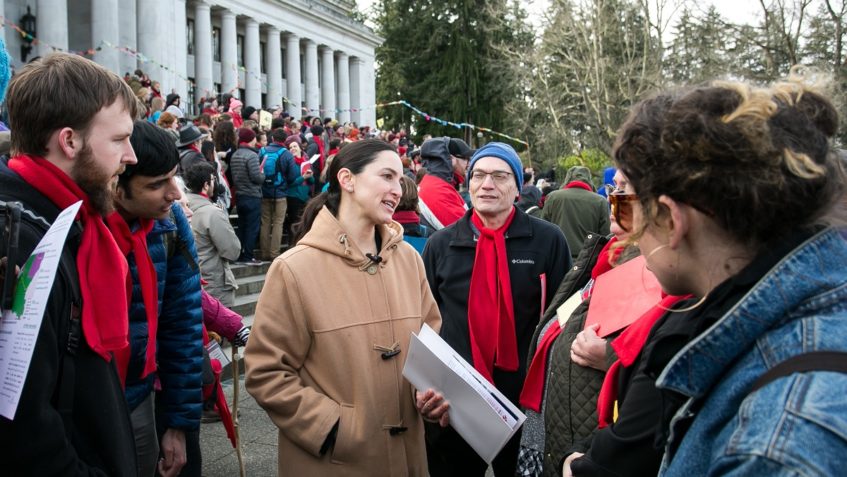 Sen. Rebecca Saldaña speaks with a a group of people standing outside the State Capitol building, and a large crowd is gathered behind them on the building's steps.