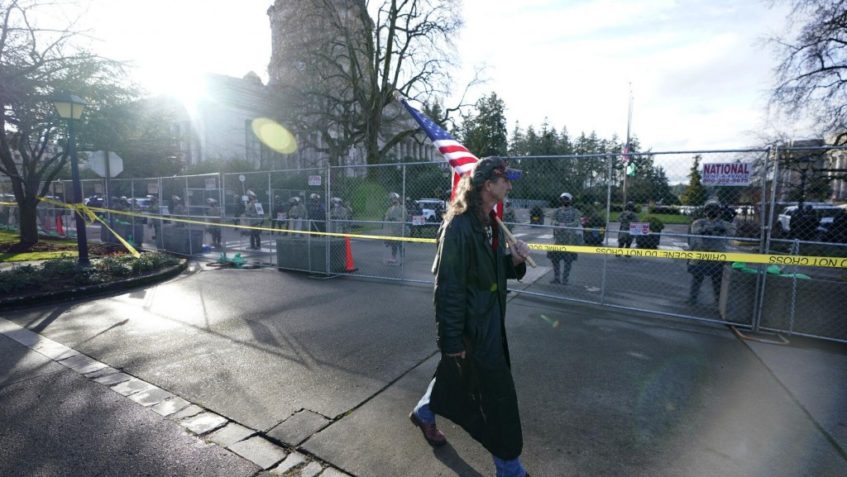 Ted S. Warren A man carries a U.S. flag past National Guard members standing behind a perimeter fence, Sunday, Jan. 10, 2021, at the Capitol in Olympia, Wash. Washington Gov. Jay Inslee activated members of the National Guard this week to work with the Washington State Patrol to protect the Capitol campus in the event of expected protests.