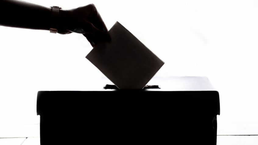 Backlit hand inserting envelope into ballot box.