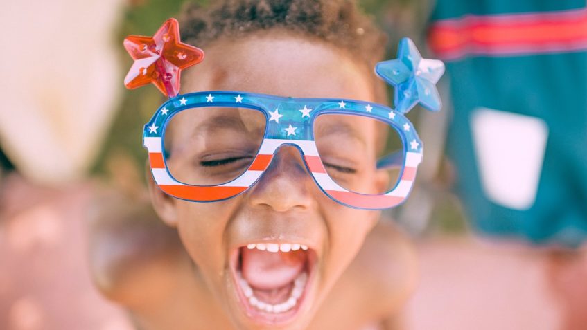 Child with mouth wide open and eyes closed wearing US flag-themed glasses with one big red star on one side, and one big blue star on the other side.