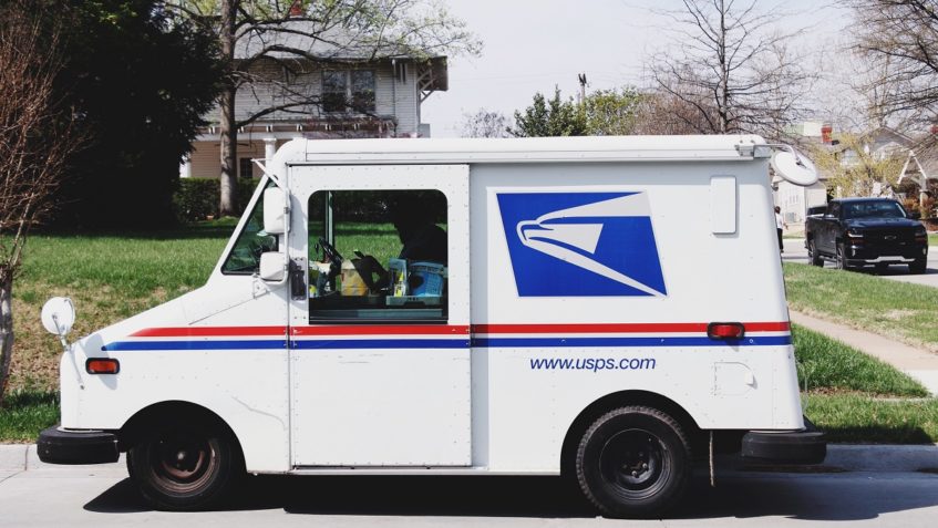 USPS mail truck parked on the street, with a house in the background. The mail carrier is seated inside the vehicle.