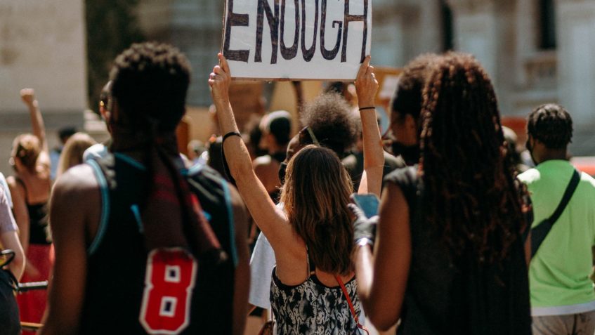 Three people march as part of a crowd. The center person is holding up a sign that says "enough."