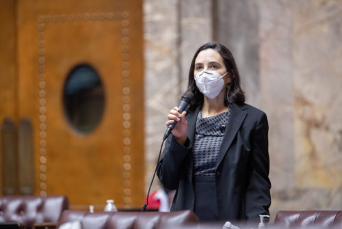 Sen. Saldaña stands at her desk on the Senate floor. She is wearing a KN-95 face mask and holding a microphone up to her face. All the seats surrounding her are empty.