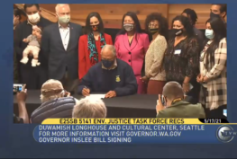Screen shot of TVW broadcast of Gov. Inslee's bill signing event for SB 5141, the Healthy Environment for All (HEAL) Act. The governor is shown seated at a table holding a paper and pen. Behind him many people are lined up to watch. In front of him, several photographers kneel taking photos.