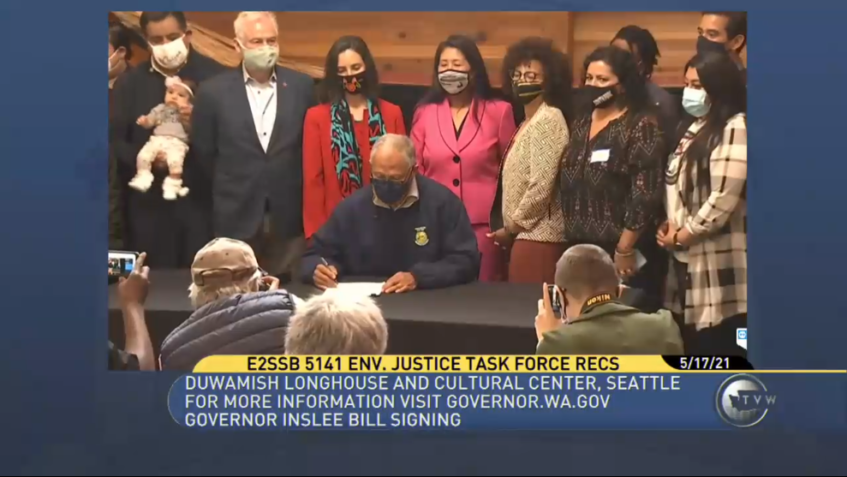 Screen shot of TVW broadcast of Gov. Inslee's bill signing event for SB 5141, the Healthy Environment for All (HEAL) Act. The governor is shown seated at a table holding a paper and pen. Behind him many people are lined up to watch. In front of him, several photographers kneel taking photos.