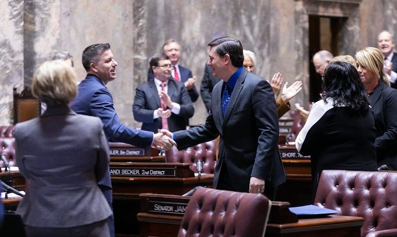 Sen. Stanford shakes hand with a fellow senator on the Senate floor while others stand and applaud.