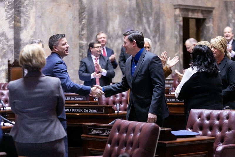 Sen. Stanford shakes hand with a fellow senator on the Senate floor while others stand and applaud.