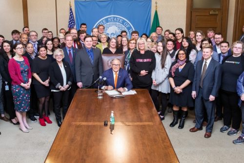 Gov. Jay Inslee is seated at the head of a long table, with a large crowd of people gathered around him, posing for a photo.