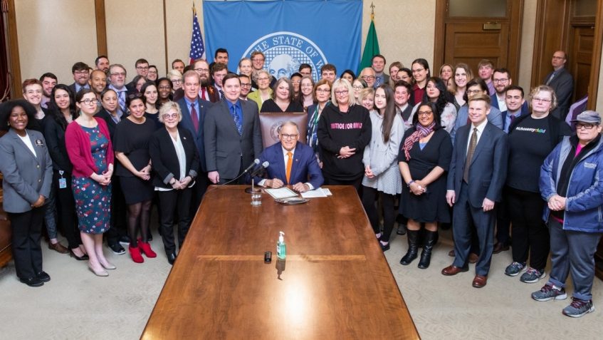 Gov. Jay Inslee is seated at the head of a long table, with a large crowd of people gathered around him, posing for a photo.