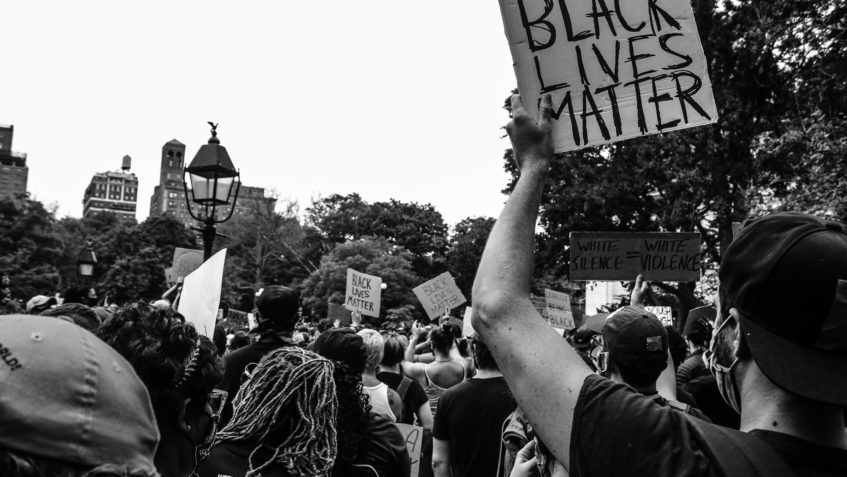 A crowd of people at a protest shown from behind. In the foreground a man holds up a sign that says "Black Lives Matter."