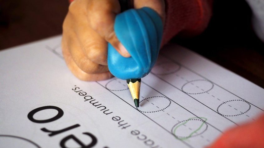 A child's hand holds a pencil in a large blue pencil grip aid, while practicing tracing zeros on a workbook page.