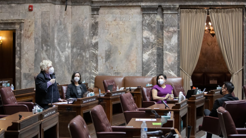 Sen. Wilson speaks while at her desk on the mostly empty Senate floor, holding a microphone. Three other senators sit at a distance at their desks, and everyone is wearing N95 face masks.