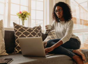 Photo of a woman sitting on a couch with an open laptop, a mug in one hand.