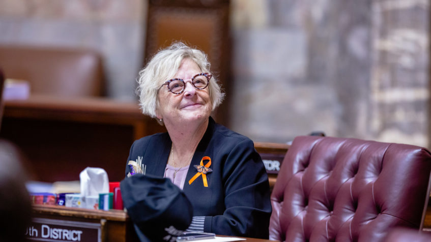 Sen. Wilson sits at her desk on the Senate floor. She's looking up toward the gallery and smiling.